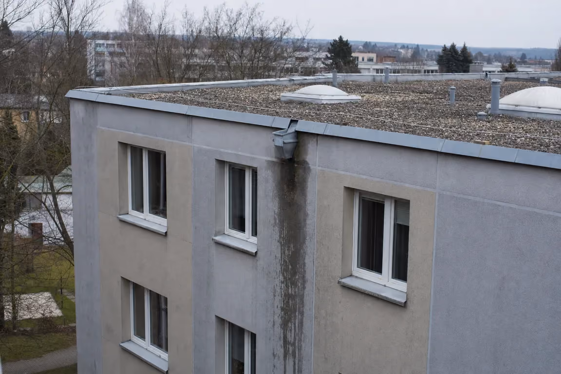 Traufbereich eines Mehrfamilienhauses nach Regen: eine dunkle Feuchtespur zieht sich von einem Rinnenauslauf über die graue Putzfassade nach unten, darüber Attikablech und Kiesdach