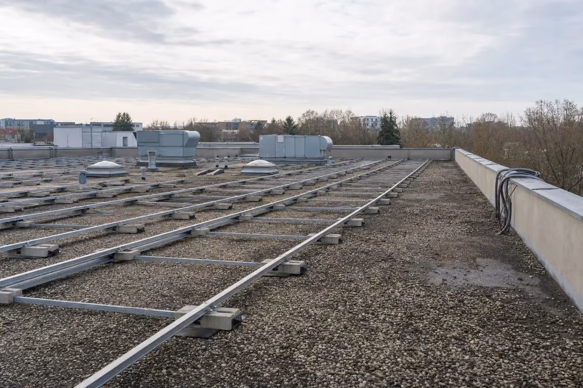 Leere Aluminium-Montageschienen auf Betonballast-Schuhen über einer Kiesdachfläche, ohne Photovoltaikmodule, aufgerollte Kabel an der Attika