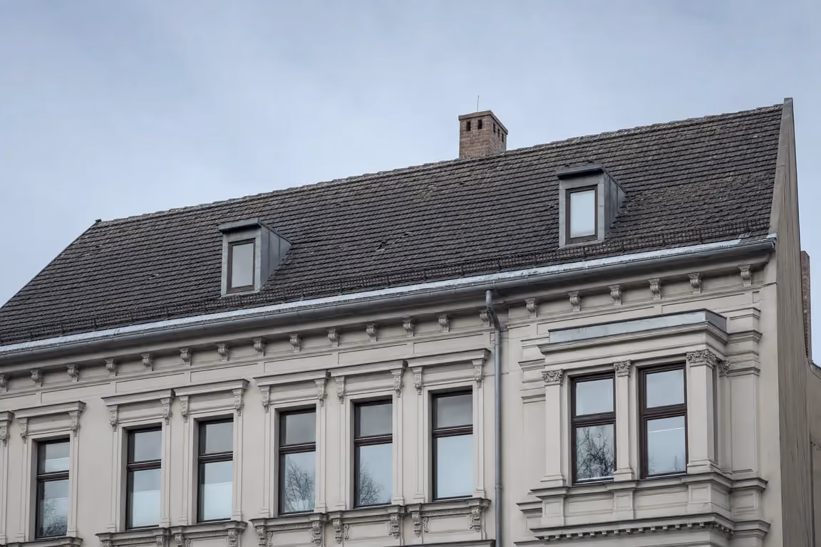 Gründerzeitliches Mehrfamilienhaus mit Stuckfassade und Erker; darüber die Traufe mit Rinne und Fallrohr und die geneigte, dunkel eingedeckte Dachfläche mit zwei Gauben und Schornstein