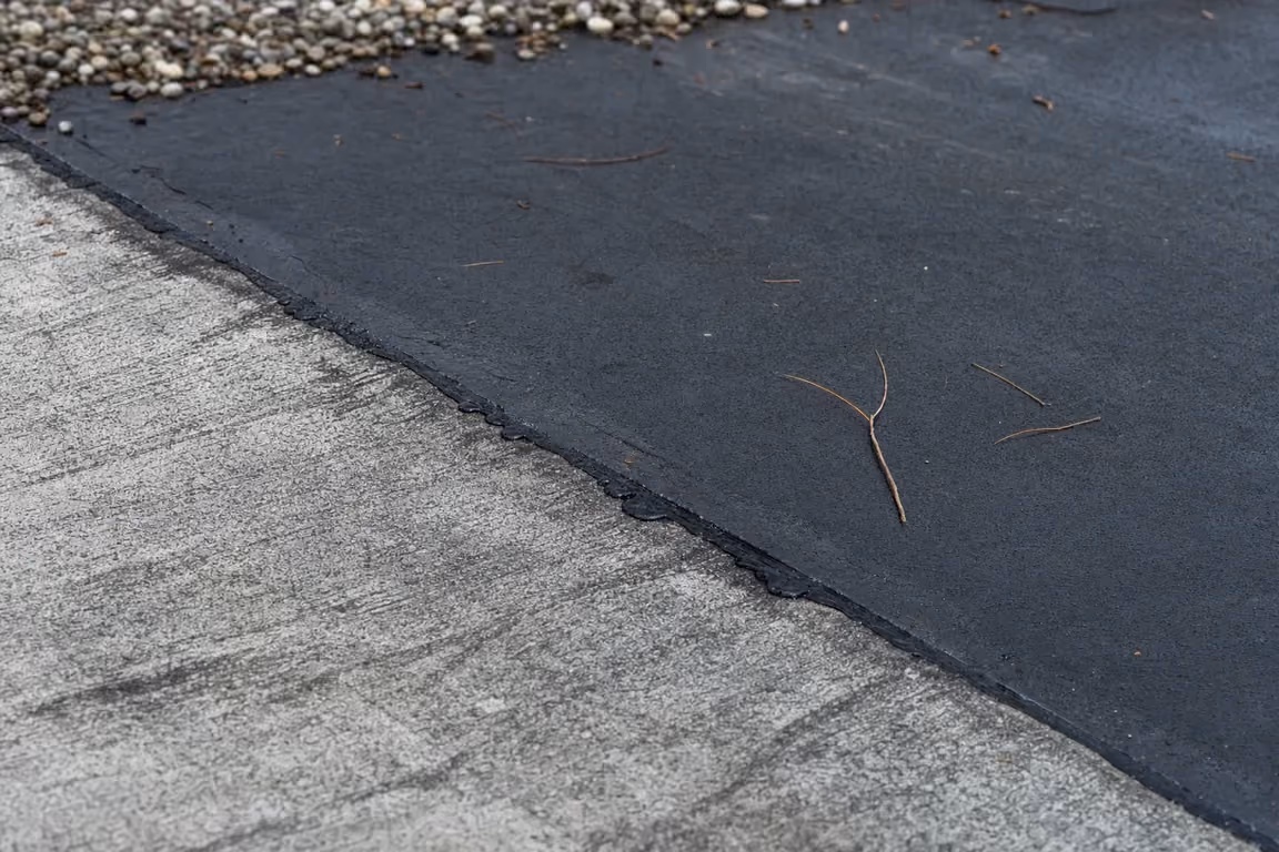 Frisch verschweißte dunkle Bitumenbahn überlappt eine grau ausgekreidete Altbahn, an der diagonalen Naht steht ausgetretene Bitumenmasse