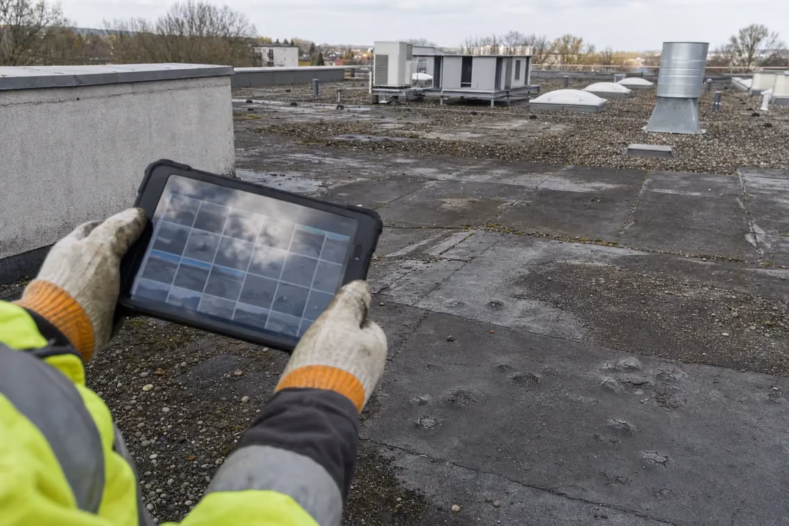 Wenn die WEG die Dachsanierung verweigert: Tablet mit der Foto-Dokumentation des Dachzustands über der gealterten, blasigen Bitumenfläche des Flachdachs